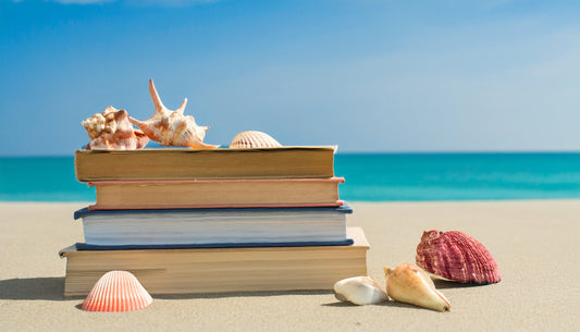 Books stacked up on a beach with seashells on top and around the books, blue ocean in the background