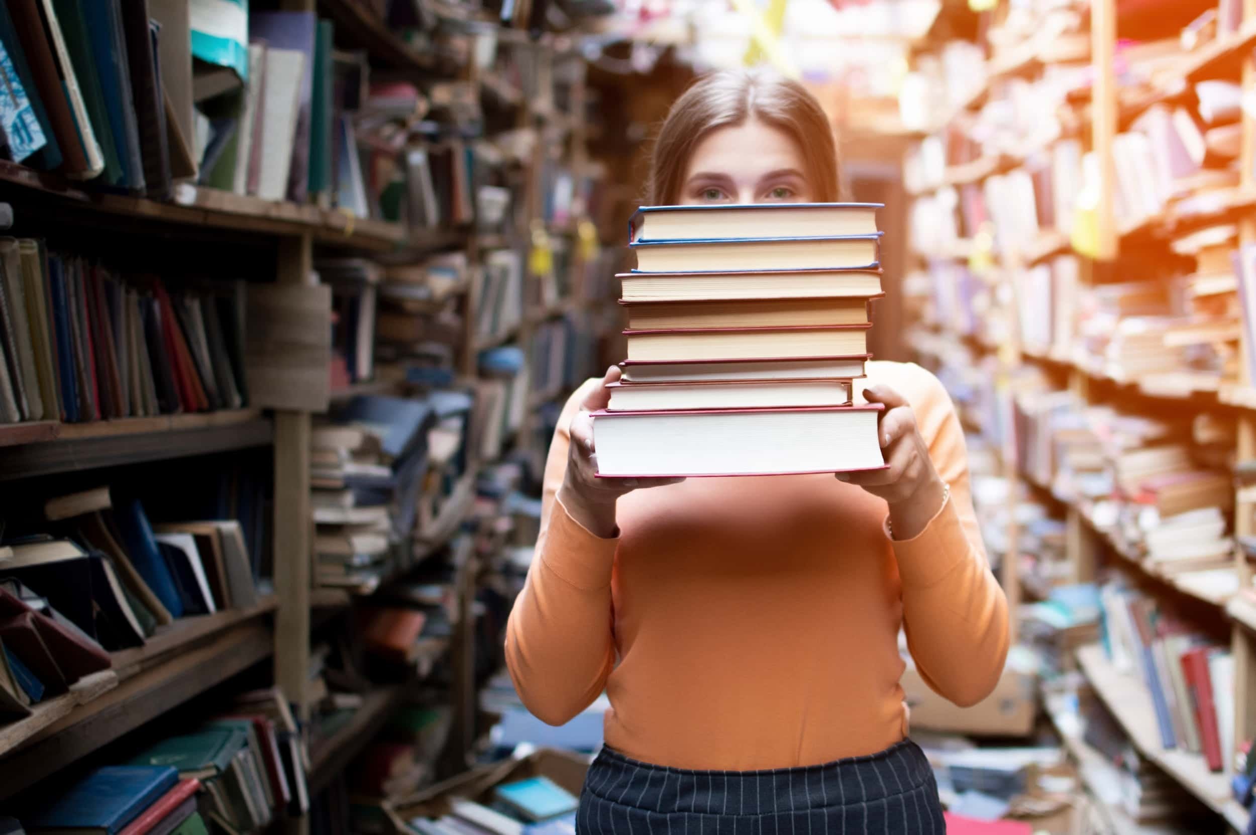 Young woman holding stack of books that is covering her face in a bookstore